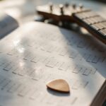 Manche de guitare et médiator posés sur un cahier de partitions ouvert